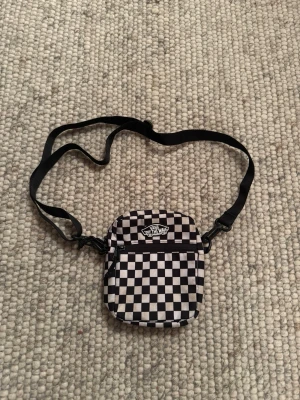 Svartvit axelväska från Vans - Liten axelväska från Vans med klassiskt svartvitt schackrutigt mönster. Väskan har justerbar axelrem, dragkedja framtill och Vans-logga på framsidan. Perfekt för att bära det viktigaste och ger en cool streetwear-vibe.