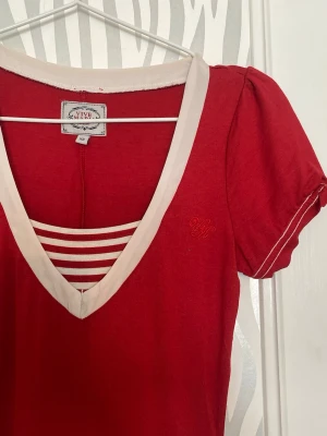 Röd klänning med v-ringning Vive Maria - Röd klänning från Vive Maria med ljus v-ringad kant och dekorativa ränder över bröstet. Puffärm och kontrastsömmar ger en söt och ungdomlig look. Klänningen har en broderad detalj på bröstet och är tillverkad i mjukt material.