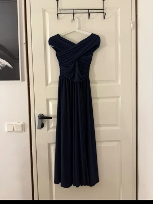 Marinblå långklänning  - Elegant marinblå långklänning med omlott över bysten och vackra draperingar. Klänningen har offshoulder-design och ett mjukt, flytande tyg som ger en snygg siluett. 