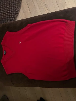 Röd v-ringad väst från GANT - Klassisk röd väst från GANT med v-ringning och ribbade kanter. Västen har en diskret logga broderad på bröstet och är tillverkad i mjukt bomullsmaterial. Perfekt att bära över en skjorta för en stilren look.