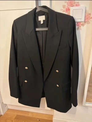 Svart dubbelknäppt kavaj H&M - Stilren svart kavaj från H&M med dubbelknäppning och silvriga knappar. Klassisk passform med breda slag och tre fickor framtill. Perfekt för en dressad look och enkel att matcha med olika outfits.