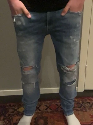 Blå jeans från Replay - Säljer ett par blå Replay Anbass jeans med slitna detaljer och en cool, avslappnad look. De har en knappgylf och klassisk femficksdesign. Perfekta för en trendig stil! Priset kan diskuteras 