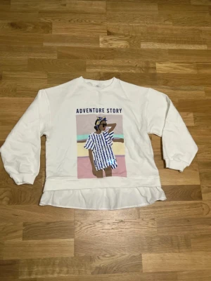 Vit sweatshirt med tryck Adventure Story - Vit sweatshirt med trycket 'ADVENTURE STORY' och motiv av en person i randig klänning på framsidan. Tröjan har långa ärmar, rund hals och en volangdetalj nedtill. Perfekt för en avslappnad och trendig look.