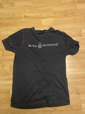 Mörkgrå Sail Racing t-shirt - Snygg mörkgrå t-shirt från Sail Racing med tryckt logga på bröstet. Klassisk passform och rund halsringning, perfekt för en avslappnad stil. Tillverkad i mjuk bomull som känns skön mot huden.