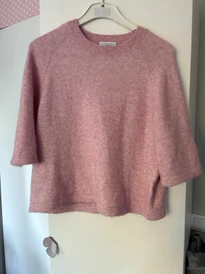 Rosa stickad tröja med halvärm - Mysig rosa stickad tröja med boxy passform och halvärm. Tröjan har rund halsringning och är tillverkad i ett mjukt, fluffigt material som ger en avslappnad look. Perfekt för lager på lager och enkel att styla till jeans eller kjol.