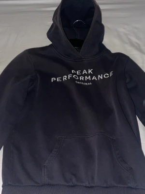 Svart hoodie Peak Performance - Snygg svart hoodie från Peak Performance med vit logga och texten 'ORIGINAL' på bröstet. Klassisk känguruficka framtill och stor huva. Perfekt för en avslappnad och sportig stil. Mjuk bomullsblandning som känns skön mot huden.