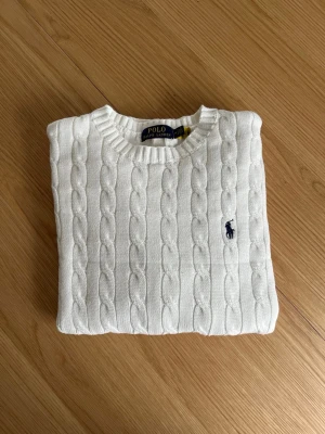 Vit kabelstickad tröja Polo Ralph Lauren - Kabelstickad vit tröja från Polo Ralph Lauren med rund halsringning och ribbade muddar. Tröjan har det klassiska broderade logotypen i marinblått på bröstet. Tillverkad i mjukt material som ger en clean och tidlös look.