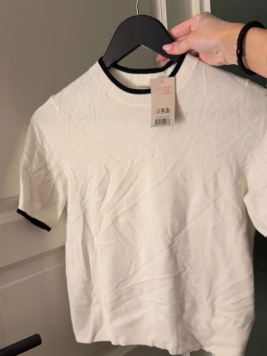 Vit kortärmad topp med svarta detaljer - Säljer en vit kortärmad topp med svarta ribbade kanter vid halsringning och ärmslut. Enkel och stilren design som passar till det mesta. Mjuk och bekväm, perfekt för dig som gillar minimalistisk stil.