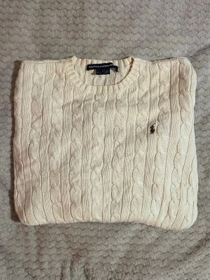 Ralph Lauren - Kabelstickad tröja i offwhite från Ralph Lauren med rund halsringning och diskret broderad logga på bröstet. Tröjan har långa ärmar och ribbade muddar vid ärmslut och nederkant. Perfekt för en klassisk och stilren look. Längd 54cm & bredd 40cm 