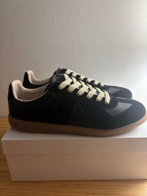 Maison Margiela gats  - Snygga svarta sneakers med beige sula och kontrasterande vita skosnören. Ovandelen är i en mix av mocka och slätt läder, vilket ger en stilren och klassisk look. Perfekta för dig som gillar minimalistisk design och neutrala färger.