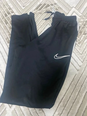 Svarta Nike mjukisbyxor med logga - Säljer ett par svarta mjukisbyxor från Nike med vit logga på låret. Byxorna har resår i midjan och smal passform. Perfekta för chill eller träning. Materialet är mjukt och stretchigt för extra komfort. Träningsbyxor  lite håll på knät