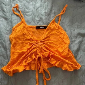 Orange croppad linnetopp från Bik Bok - Säljer en orange croppad linnetopp från Bik Bok med smala axelband, v-ringning och snygg snörning framtill. Toppen har volangkant nedtill och är perfekt för sommardagar. Materialet känns lätt och luftigt.