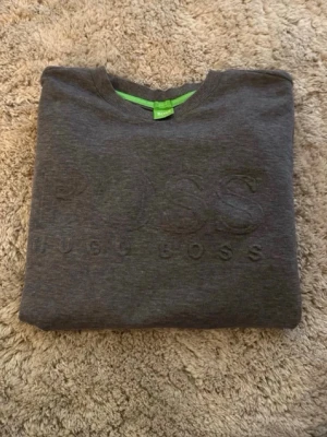 Mörkgrå sweatshirt från BOSS - Snygg mörkgrå sweatshirt från BOSS med diskret broderad logga framtill. Tröjan har rund halsringning och ribbade muddar. Perfekt för en clean och stilren look. Materialet känns mjukt och bekvämt mot huden.