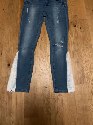 Blå jeans med spetsdetaljer - Unika blå jeans med slitningar och vita spetskilar längs bensluten. Jeansen har en klassisk femficksmodell och raka ben med extra vidd tack vare spetsen. Perfekta för dig som vill sticka ut med en kreativ och trendig look. Innerbenslängd 71 midjemått 40, storlek 30/30💞