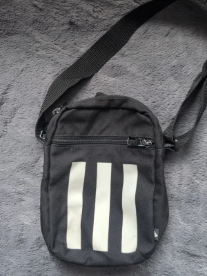 Adidas axelremsväska svart  - Säljer en svart axelväska från Adidas med tre vita ränder på framsidan. Väskan har justerbar axelrem, dragkedja upptill och ett mindre ytterfack med dragkedja. Tillverkad i slitstark textil, perfekt för att bära det viktigaste.