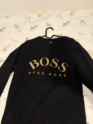 Svart sweatshirt från Hugo Boss - Snygg svart sweatshirt från Hugo Boss med stort guldfärgat BOSS-tryck på bröstet. Tröjan har rund halsringning och ribbade muddar vid ärmslut och nederkant. Perfekt för en stilren och trendig look.