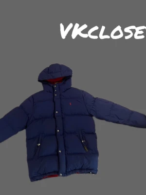 Marinblå dunjacka från Polo Ralph Lauren - Mörkblå dunjacka med huva från Polo Ralph Lauren. Jackan har röd insida, två framfickor med dragkedja och tryckknappar framtill. Klassisk puffer-modell med diskret logga på bröstet. Perfekt för kalla vinterdagar. Storlek L(14/16)