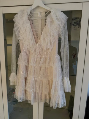 Vit spetsklänning med volanger - Romantisk kort vit klänning i spets med djup v-ringning, långa ärmar och flera lager volanger. Transparenta spetsdetaljer och pärlknappar framtill ger en extra elegant känsla. Perfekt för dig som gillar en feminin och unik stil.