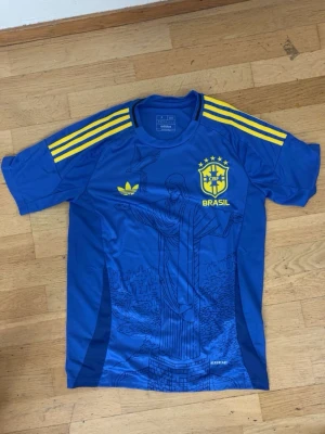 Brasilien fotbollströja - Snygg blå fotbollströja från Adidas med Brasilien-tryck och gula detaljer. Tröjan har gula ränder på axlarna, CBF-logga och Adidas-logga på bröstet samt ett stort tryck av Kristusstatyn på framsidan. Perfekt för dig som älskar fotboll och vill sticka ut.