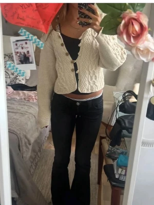 Vit kabelstickad kofta med knappar - Säljer en krämvit kabelstickad kofta med snygga träknappar framtill. Koftan har en croppad passform, lång ärm och ett klassiskt flätmönster både fram och bak. Perfekt att slänga över ett linne för en chill look.