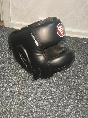 Svart boxningshjälm från RDX - Säljer en svart boxningshjälm från RDX, modell HGR T8. Hjälmen är tillverkad i slitstarkt konstläder och har vaddering runt hela huvudet samt extra skydd för hakan. Perfekt för sparring och träning inom kampsport.