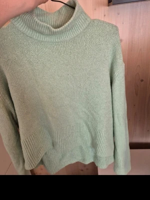 Mintgrön stickad polotröja - Mysig mintgrön stickad tröja med hög polokrage och ribbade detaljer vid hals, ärmslut och nederkant. Tröjan har en avslappnad passform och är perfekt för kyliga dagar. Materialet känns mjukt och behagligt mot huden.