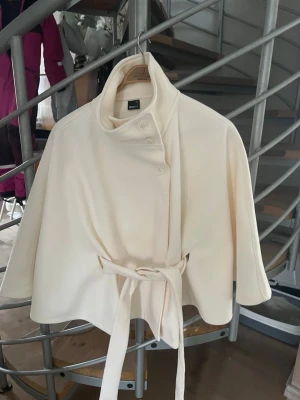Gina poncho - Stilren krämvit cape-kappa från gina med hög krage, knappar framtill och knytband i midjan. Kappan har vida trekvartsärmar och en mjuk, slät yta som ger en elegant look. Perfekt för dig som vill ha en trendig och unik ytterjacka.