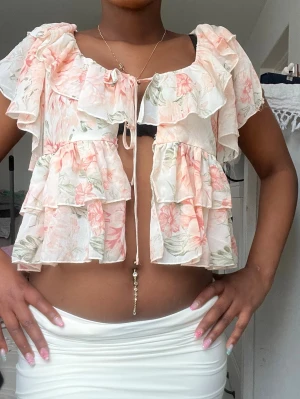 Blommig volangblus med knyt - Supersöt blommig blus med volanger och knyt framtill. Luftigt material i ljusa toner av rosa, grön och vit. Korta ärmar och öppen framtill för en trendig look. Perfekt för vår och sommar när du vill känna dig extra fin.