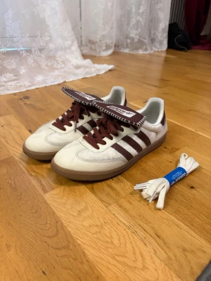 Adidas Wales Bonner Samba sneakers - Snygga Adidas Samba sneakers i samarbete med Wales Bonner. Skorna är vita med bruna detaljer, bruna ränder och matchande bruna skosnören. Ovandelen är i läder och mocka, med gummisula och extra vita skosnören medföljer. Unik design på plösen med Wales Bonner-logga.