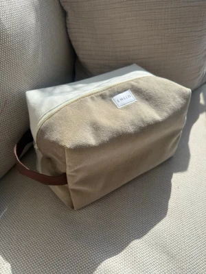 Beige necessär från Emelo - Stilren necessär i beige och vitt sammet från Emelo. Rektangulär form med dragkedja upptill och ett brunt handtag i sidan. Perfekt för att organisera smink eller småprylar. Tillverkad i sammetstyg för en mjuk och fräsch look. Mått ca 25x17x12cm