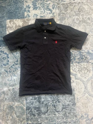 Svart pikétröja från Polo Ralph Lauren storlek M - Klassisk svart pikétröja från Polo Ralph Lauren med korta ärmar och krage. Tröjan har två knappar framtill och den ikoniska röda logotypen broderad på bröstet. Tillverkad i mjuk bomull för en bekväm känsla. Tröjan är helt ny och aldrig buren och har storlek M