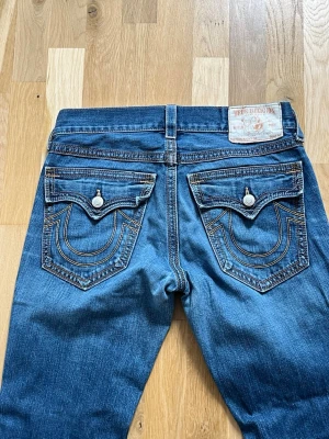 Blå True Religion jeans med kontrastsömmar - Säljer ett par blå jeans från True Religion med ikoniska kontrastsömmar och stora ficklock bak. Jeansen har en klassisk femficksmodell, knappgylf och detaljerade sömmar på bakfickorna. Materialet är denim i bomull och passformen är rak.