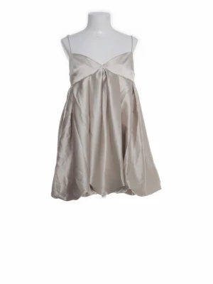 Champagnefärgad kort klänning i satin - Snygg kort klänning i champagnefärgad satin med tunna axelband och ballongliknande kjol. Klänningen har en v-ringad överdel och smock i ryggen för skön passform. Perfekt för dig som vill ha en elegant och trendig look.