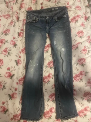 Miss me bootcut lågmidjade jeans - Säljer ett par blå bootcut jeans från Miss Me med slitningar och dekorativa bakfickor med broderi och nitar. Jeansen har låg midja, klassisk femficksmodell och kontrastsömmar. Jag vet inte om färgfläckarna var original design eller om de var förra ägaren som har gjort de för jag köpte de här på Plick och jag tycker de blir väldigt coola med de🫶🏻Jag har sytt in jeansen så de blivit mindre än storleken som det står. Jeansen är storlek 31 men de passar en M skulle jag säga💕
