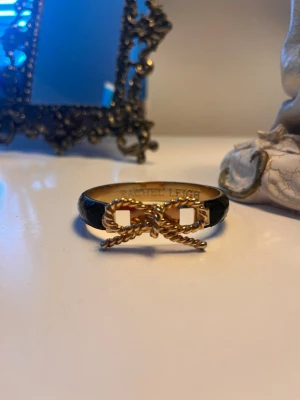 Svart armband med guldfärgad rosett - Stilrent armband från Rachel Leigh i svart med en dekorativ guldfärgad rosett framtill. Armbandet har en elegant och ungdomlig design med metallisk finish och passar perfekt för att lyfta din outfit.