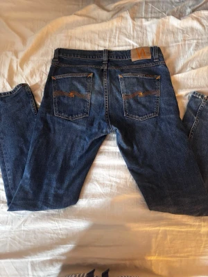 Mörkblå jeans från Nudie Jeans - Säljer ett par klassiska mörkblå jeans från Nudie Jeans med raka ben och snygga kontrastsömmar. Jeansen har fem fickor och ikoniska broderade detaljer på bakfickorna. Perfekta för en avslappnad och stilren look. Str: 30:32