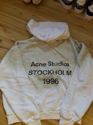 Vit hoodie Acne Studios Stockholm - Säljer en vit hoodie från Acne Studios med trycket 'Acne Studios STOCKHOLM 1996' på framsidan. Hoodien har en klassisk huva och ribbade muddar. Perfekt för dig som gillar streetwear och vill ha en stilren look.
