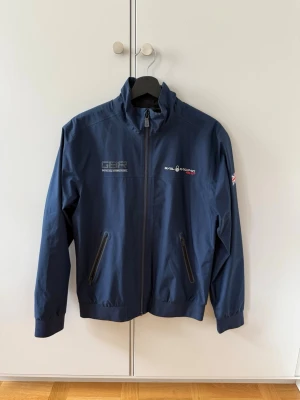 Marinblå vindjacka från Sail Racing - Snygg marinblå vindjacka från Sail Racing med hög krage och dragkedja framtill. Jackan har två snedställda fickor med dragkedja och brittisk flagga på ärmen. Tillverkad i ett lätt och vindtätt material, perfekt för blåsiga dagar.