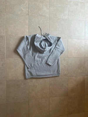 Ljusgrå hoodie med broderad detalj - Säljer en stilren ljusgrå hoodie med huva och dragsko. Tröjan har en liten broderad logga på bröstet och är tillverkad i mjukt stickat material. Perfekt för dig som gillar en clean och enkel look.