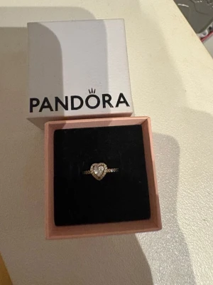 Pandora guldring med hjärtformad sten - Snygg guldfärgad ring från Pandora med en stor hjärtformad klar sten i mitten, omgiven av små gnistrande stenar längs hela ringen. Perfekt för dig som gillar eleganta och romantiska smycken. Kommer i originalask.