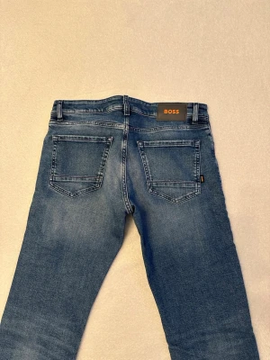 Blå jeans från BOSS med raka ben - Snygga blå jeans från BOSS med klassisk femficksmodell och raka ben. Jeansen har en ljus tvätt och subtila slitningar för en avslappnad look. Märkespatch bak i midjan och detaljerade sömmar. Perfekta för dig som gillar stilrena och tidlösa jeans. Oanvända! Nypris 1600kr
