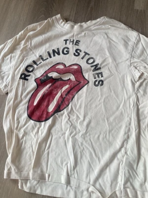 Rolling Stones t-shirt från H&M - Vit t-shirt från H&M med stort Rolling Stones-tryck framtill. Klassisk rund hals och korta ärmar. Ikonisk röd tunga och svart text ger en cool retrostil. Tillverkad i mjuk bomull som är skön att bära hela dagen.