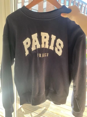 Mörkblå sweatshirt med PARIS-tryck - Mörkblå sweatshirt med vit text 'PARIS FRANCE' på bröstet. Tröjan har rund halsringning, ribbade muddar vid ärmslut och nederkant samt en avslappnad passform. Perfekt för en casual och trendig look.