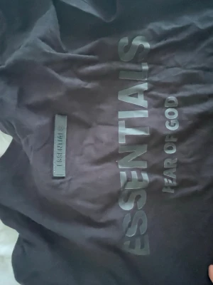 Svart Essentials Fear of God hoodie - Snygg svart hoodie från Fear of God Essentials med stor logga på bröstet och mindre logga på ryggen. Tillverkad i mjukt bomullsmaterial och har en klassisk oversized passform. Perfekt för dig som gillar streetwear och vill ha en clean look.