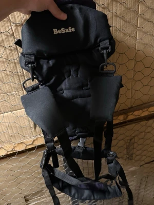 BeSafe svart och lila bärsele - Bärsele från BeSafe i svart och lila tyg med broderad detalj framtill. Selen har justerbara remmar, spännen och en praktisk ficka. Materialet är slitstarkt tyg och designen är ergonomisk för att bära barn nära kroppen.