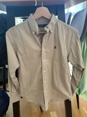 Beige skjorta från Polo Ralph Lauren - Klassisk beige skjorta från Polo Ralph Lauren med button-down krage och broderad logga på bröstet. Skjortan har lång ärm och knappar framtill. Perfekt till en avslappnad men ändå stilren look.
