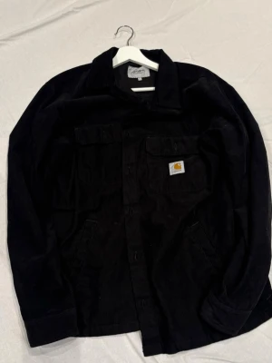 Svart overshirt från Carhartt - Säljer en svart overshirt från Carhartt i mjuk manchester. Skjortan har två bröstfickor, varav en med klassisk Carhartt-logga, samt sidofickor. Perfekt lager-på-lager-plagg med avslappnad passform och stilren look.