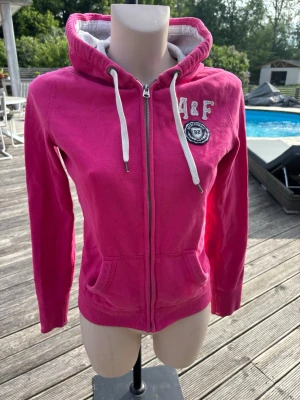 Rosa hoodie från Abercrombie & Fitch - Snygg rosa hoodie med dragkedja från Abercrombie & Fitch. Har vit dragsko i huvan, broderad logga och fickor framtill. Mjuk insida och klassisk passform, perfekt för en avslappnad stil.