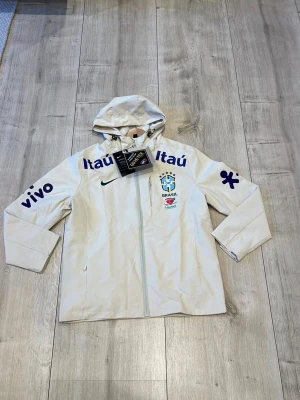 Vit Nike Brasil vindjacka med huva - Snygg vit vindjacka från Nike med Brasilien CBF-logga och flera sponsortryck i blått. Jackan har huva, dragkedja framtill och är tillverkad i slitstarkt GORE-TEX-material. Perfekt för dig som gillar sportig stil och vill sticka ut.