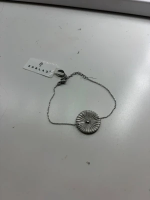 Silverfärgat armband från Edblad - Snyggt armband från Edblad i silverfärg med en tunn kedja och ett runt, räfflat hänge med en liten sten i mitten. Perfekt accessoar för dig som gillar stilrena och moderna smycken. Lätt att matcha med andra armband eller klockor.